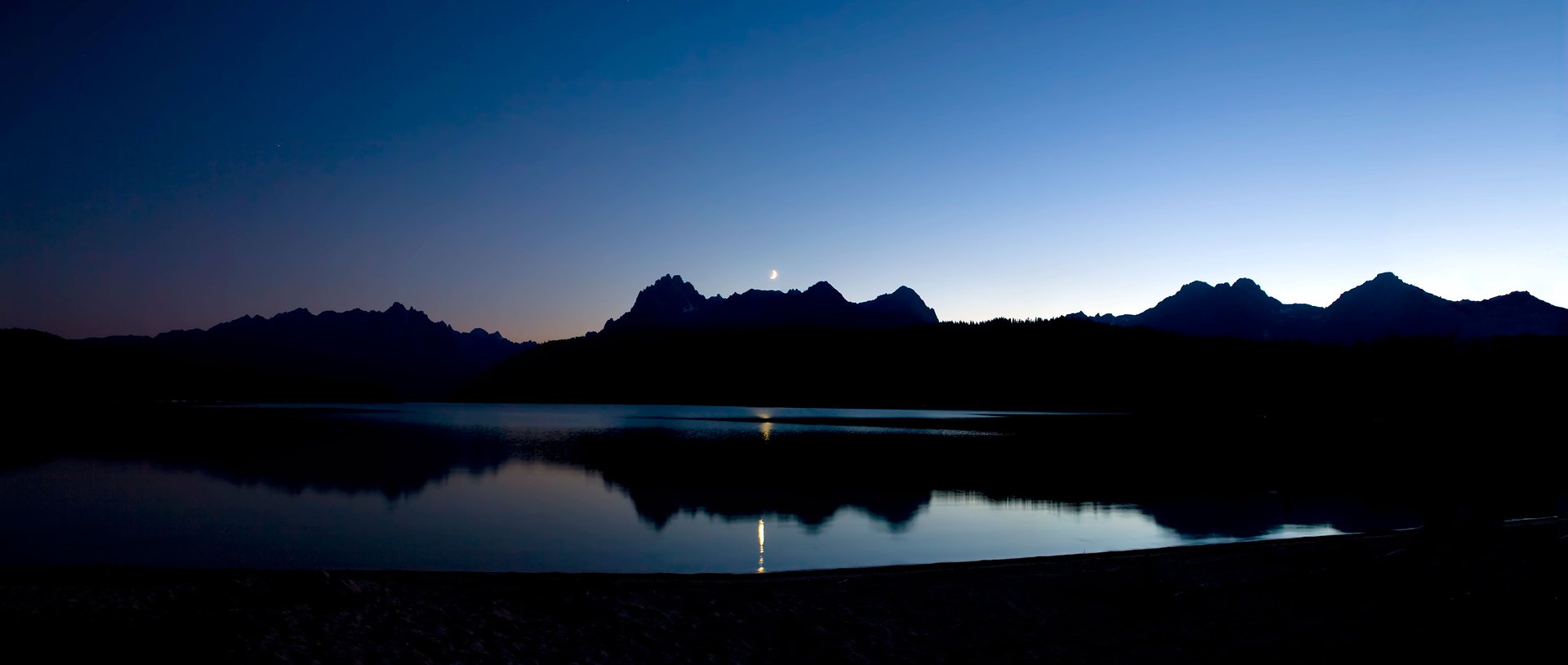 Night panorama of Redfish Lake, Idaho. The jagged Sawtooth peaks stand in black silhouette against a deep blue sky fading to violet at the horizon. A thin crescent moon hangs just above the central peaks, and its light falls in a narrow silver streak across the still, mirror-like lake toward the dark shoreline.